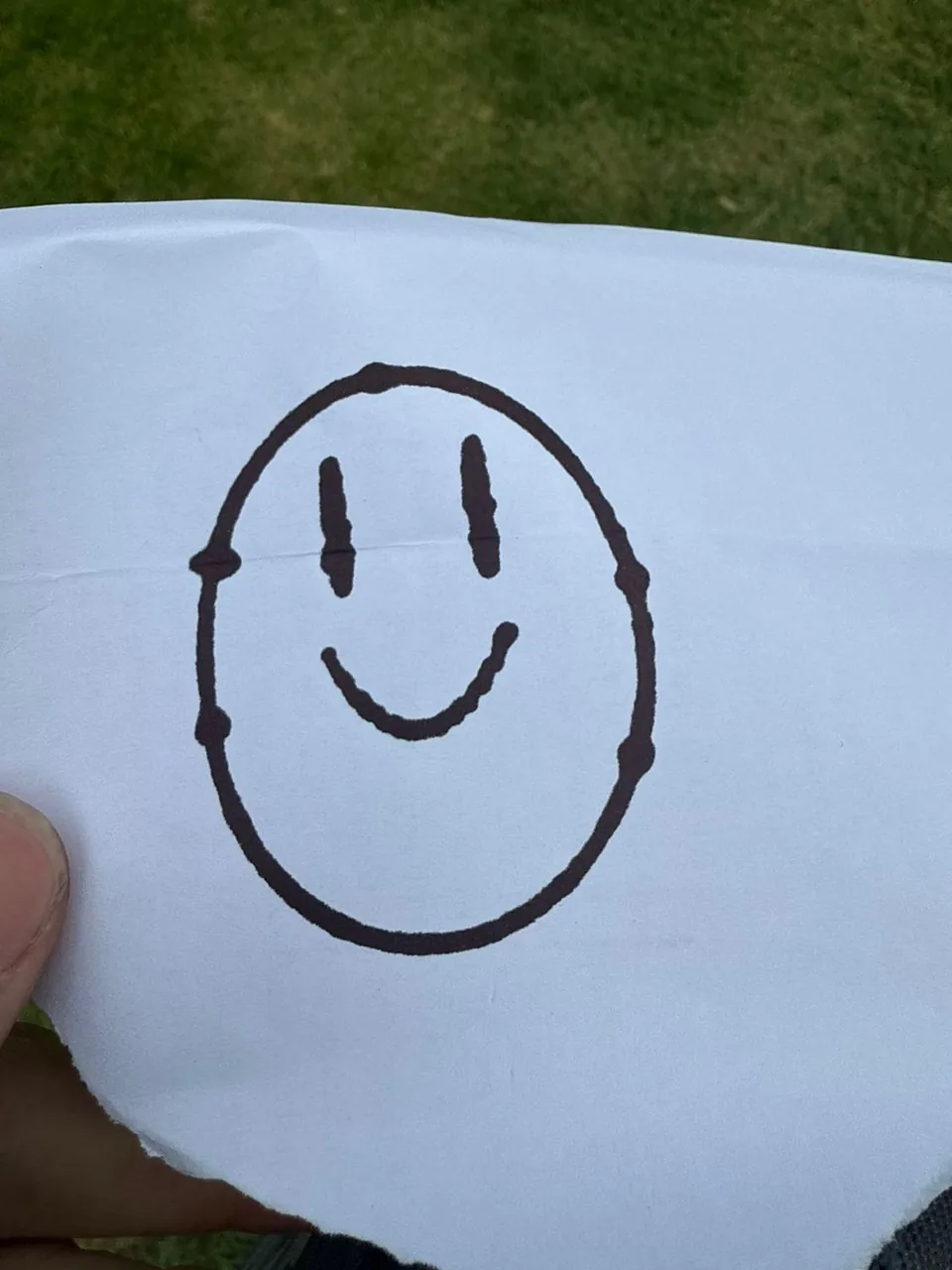 A smiley face drawn by the machine on a torn sheet of paper, held up outdoors.