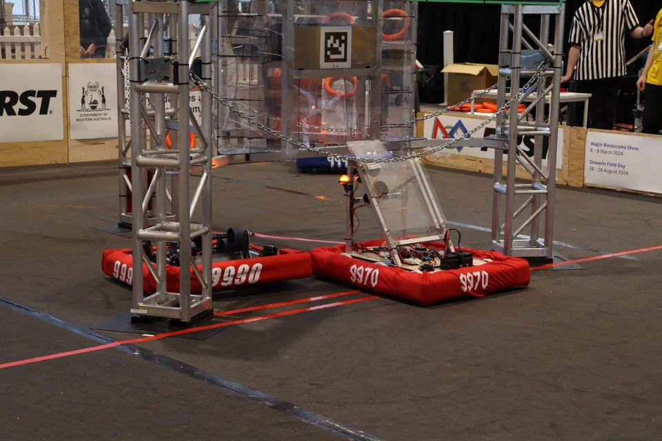 Red-bumpered robots on the field beneath a metal truss, with FIRST banners behind them.